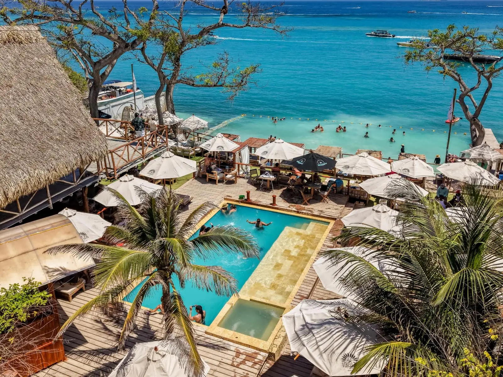 Vista aérea de Ibbiza Island Beach Club: piscina, sombrillas blancas, palmeras y el mar turquesa con bañistas y botes