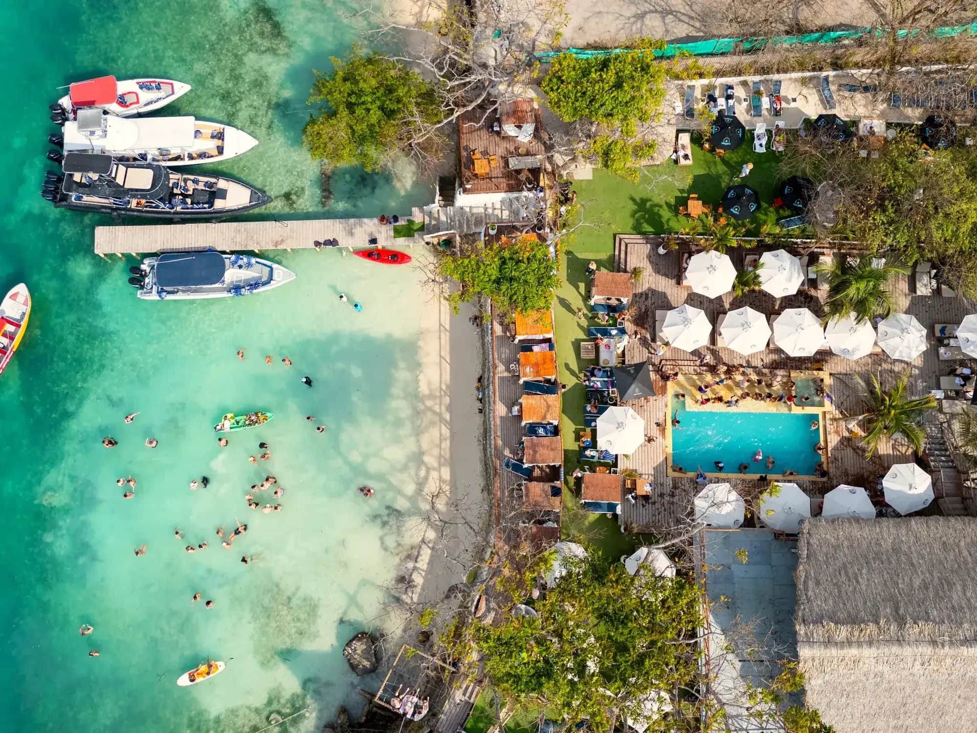 Vista aérea de Ibbiza Island Beach Club: piscina, sombrillas blancas, muelle con botes y bañistas en el mar turquesa